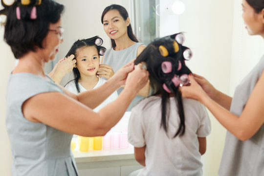 Mother and grandmother curling curlers to little girl together in front of the mirror and preparing for holiday event