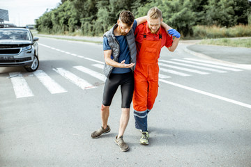 Medic in uniform helping injured man to walk, applying first aid after the road accident