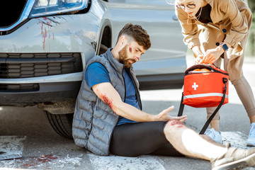 Driver hurrying with first aid kit to help injured man with bleeding wounds sitting near the car...