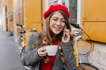 Pleased young woman in gray jacket talking on phone and drinking coffee in street cafe. Outdoor portrait of blissful french girl in red beret calling friend, while enjoying hot beverage in cold day.