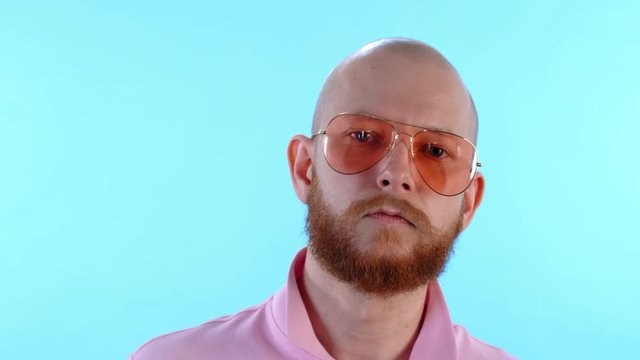 Close up face of ginger haired bearded man putting on stylish sunglasses and looking at camera on light blue background