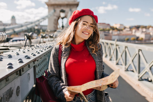 Outdoor photo of pretty female traveler in glasses holding map on the city bridge. Attractive european woman in red sweater and beret exploring country in autumn vacation.