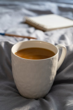 Hygge, lagom lifestyle image with coffee cup, open notebook and a pencil on the bed, covered with minimalist grey linen sheet. Cozy lifestyle.