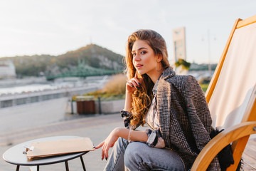 Attractive young woman in vintage jeans sitting on recliner in outdoor restaurant. Lovable brunette girl in tweed coat waiting for order in favorite cafe and enjoying nature views.