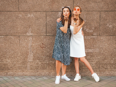 Two young beautiful smiling hipster girls in trendy summer dresses. Sexy carefree women posing in the street near brown wall.Positive models having fun in sunglasses.Giving air kiss