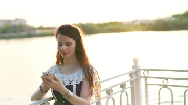 Close-up portrait of a woman girl with brown hair in a sundress in the summer at sunset on the waterfront using a smartphone cell phone on the Internet in social networks