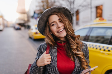 Close-up portrait of refined young lady with nude make-up smiling while posing on avenue in...