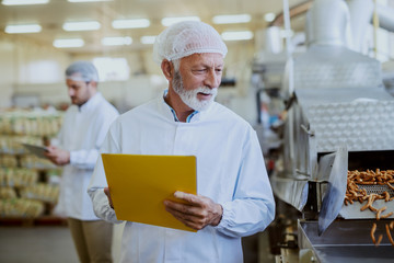 Fototapeta na wymiar Caucasian senior adult quality controller holding folder with documents and checking on quality of salty sticks. Food plant interior.