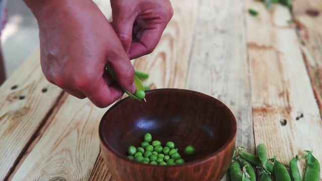 closeup of woman hand depod green peas in brown dish, on wooden table.