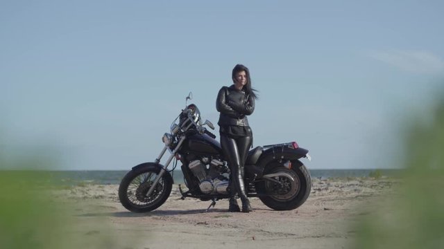 Attractive brunette caucasian girl in leather clothes standing near the motorcycle at the beach of the sea. Hobby, traveling and active lifestyle. Leisure and travel by motorbike.