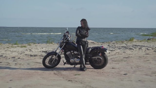 Cute brunette caucasian girl in black leather clothes standing near the motorcycle at the beach of the sea. Hobby, traveling and active lifestyle. Leisure and travel by motorbike.