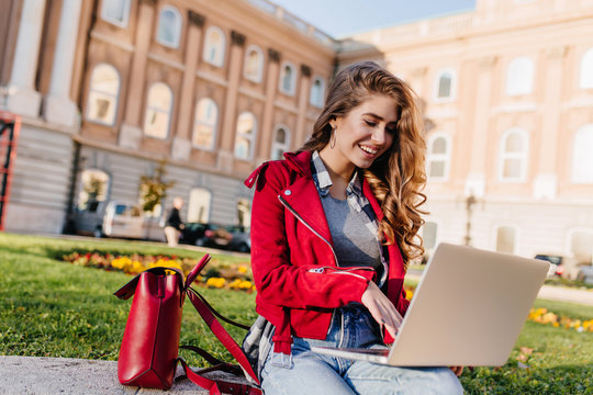 Laughing female student with leather backpack typing on laptop keyboard while chiling beside university. Smiling freelancer in red jacket working with computer, sitting on grass.