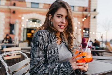 Joyful dark-haired girl in tweed jacket holding glass of cocktail. Pretty white female model...