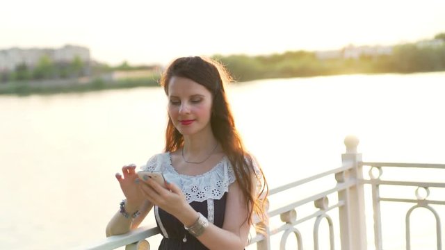 Close-up portrait of a European woman girl with brown hair in a sundress in the summer at sunset on the waterfront using a smartphone cell phone on the Internet in social networks