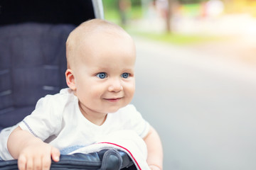 Cute caucasian blond curious baby boy sitting in stroller , smiling, looking and aspiring to something outdoor at city park. Little toddler child having fun walking in pram with family at nature