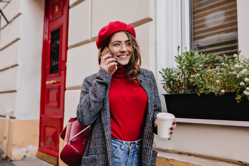 Winsome girl in stylish casual attire talking on phone, walking past cafe. Outdoor portrait of cute...