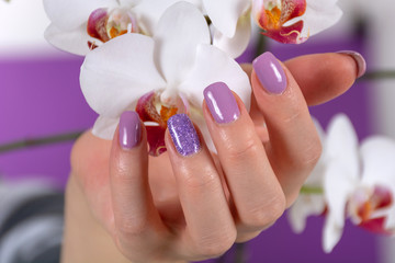 Female hand with a lilac color nails polish gel and beautiful orchid flower decoration on purple background in the studio. Manicure and beauty concept. Close up, selective focus