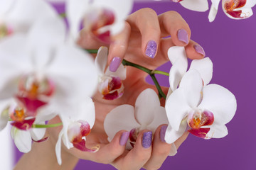 Young female hand with a lilac color nails polish gel and beautiful orchid flower decoration on purple background in the studio. Manicure and beauty concept. Close up, selective focus