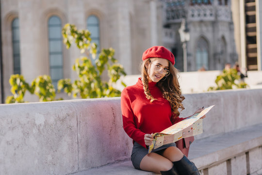Winsome girl in short skirt sitting on stone bench with city map. Attractive french lady with curly hairstyle posing on old architecture background.