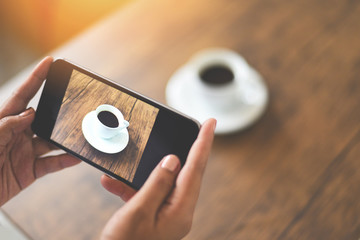 Taking photo coffee cup with camera smartphone for use technology post upload file to online on Internet - Close up of woman hand holding mobile phone photographing coffee cup on wooden table