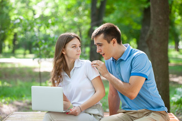 Young couple sitting together on bench in park and using laptop