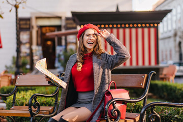 Pleased girl in short skirt sitting in cozy city square and laughing. Lovable french lady in gray jacket posing with newspaper on bench.