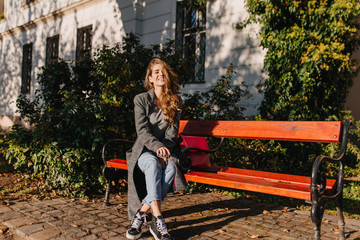 Inspired girl in long coat sitting on wooden bench near her house early in evening. Outdoor portrait of blonde woman in jeans resting beside green trees in sunny october day.