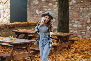 Slim girl in blue denim pants talking on phone in park with trees behind. Outdoor photo of...