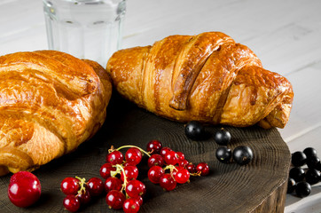 Fresh ruddy croissants with berries lie on a wooden table next to fresh black currant berries, red currants, cherries