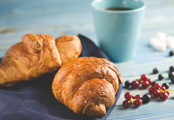 Fresh ruddy croissants with berries lie on a wooden table next to fresh black currant berries, red currants, cherries