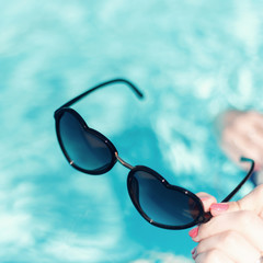 Women's hand with a beautiful manicure holding sunglasses in shape of heart on a background against the azure water of the pool. The concept of summer, love and relaxation. Fashion summer accessory