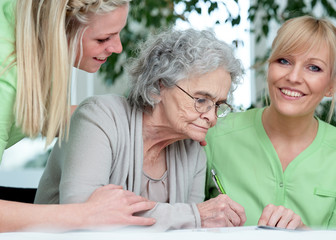 Pflegerinnen helfen Seniorin im Rollstuhl beim Kreuzworträtzeln