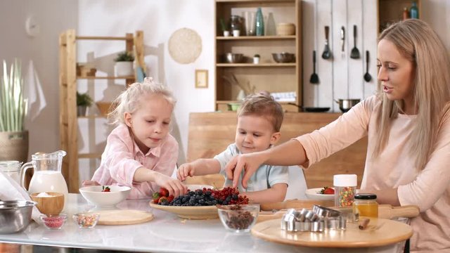 Waist-up shot of young Caucasian mother with two kids sitting at kitchen table, with baking utensils, condiments and ingredients on it, and sorting plate of fresh strawberries, blueberries and currant