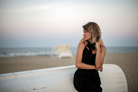 Beautiful woman in black summer dress on the beach near white wooden boat.