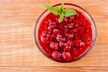 Glass small saucer with raspberry jam with fresh raspberries and mint close-up on wooden background.