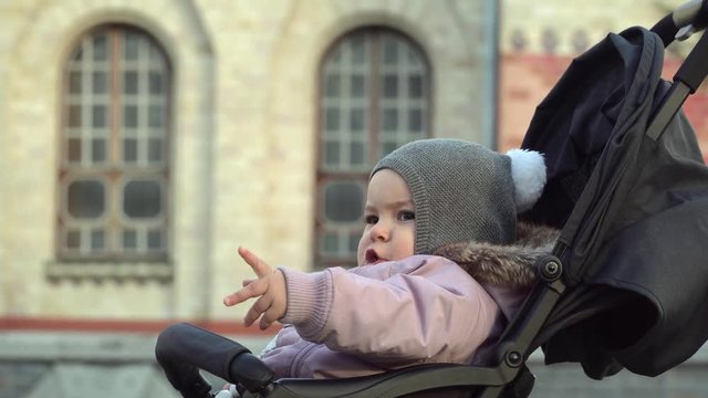 Portrait of one year old girl sits in stroller. Close up of travelling baby. Cold weather. 4k