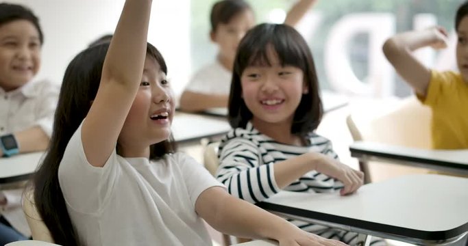 Group of joyful asian elementary students raising their arms to answer a question from teacher at classroom. Kindergarten pre school concept.