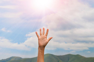 a woman's hand reaches up to the sunlight with rays against the blue sky with clouds
