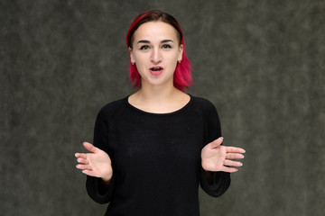 Portrait to the waist of a pretty girl with red hair on a gray background in a black jacket. Standing right in front of the camera in a studio with emotions, talking, showing hands, smiling