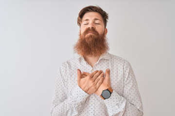 Young redhead irish man wearing shirt standing over isolated white background smiling with hands on chest with closed eyes and grateful gesture on face. Health concept.