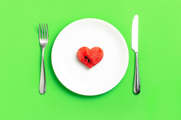 Creative summer food diet concept. Juicy slices of ripe red watermelon in the shape of a heart in white plate, knife, fork on green background. Flat lay top view. Love for watermelon