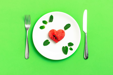 Creative summer food concept. Juicy slices of ripe red watermelon in the shape of a heart and mint leaves in white plate, knife, fork on green background. Flat lay top view. Love for watermelon