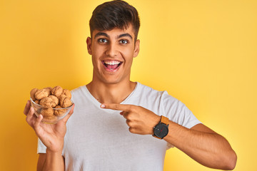 Young indian man holding bowl with walnuts standing over isolated yellow background very happy pointing with hand and finger