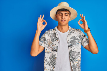Indian man on vacation wearing hawiaian shirt summer hat over isolated blue background relax and smiling with eyes closed doing meditation gesture with fingers. Yoga concept.