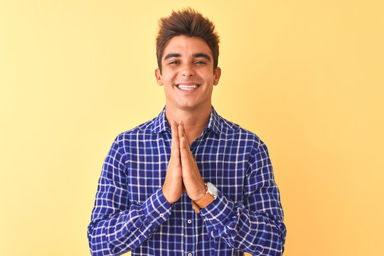 Young handsome man wearing casual shirt standing over isolated yellow background praying with hands together asking for forgiveness smiling confident.