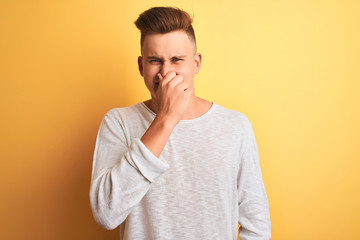 Young handsome man wearing white casual t-shirt standing over isolated yellow background smelling something stinky and disgusting, intolerable smell, holding breath with fingers on nose. 