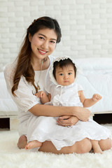 Fototapeta na wymiar Happy Beauty mother Holding and kissing Cute Sweet Adorable Asian Baby wearing white dress sitting on Carpet smiling and playing with happiness emotional in cozy bedroom,Healthy Baby Concept