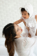 Fototapeta na wymiar Happy Beauty mother Holding Cute Sweet Adorable Asian Baby wearing white dress smiling and playing with happiness emotional in cozy bedroom,Healthy Baby Concept