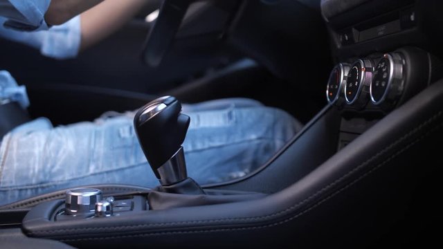Close-up of female driver hand switching automatic gearbox in modern expensive car while driving. Casually dressed woman sitting on driver's seat shifting gear knob driving automobile, interior view