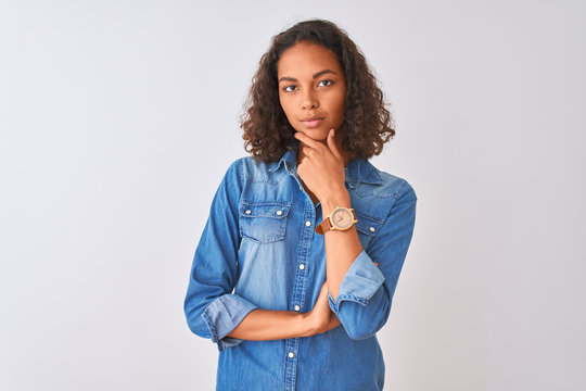 Young brazilian woman wearing denim shirt standing over isolated white background looking confident at the camera smiling with crossed arms and hand raised on chin. Thinking positive.